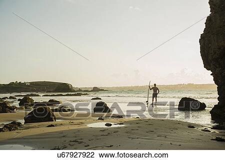 Stock Image - Mature man standing at waters edge holding surf board. Fotosearch