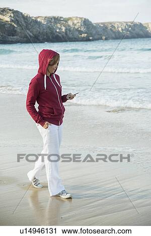 Stock Image - Mature woman reading text message on smartphone whilst strolling on beach, Camaret-sur-mer, Brittany, France. Fotosearch