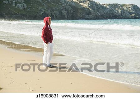 Stock Image - Mature woman standing on beach with hands in pockets, Camaret-sur-mer, Brittany, France. Fotosearch
