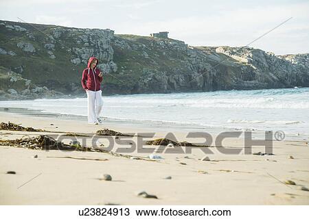 Stock Image - Mature woman texting on smartphone whilst strolling on beach, Camaret-sur-mer, Brittany, France. Fotosearch