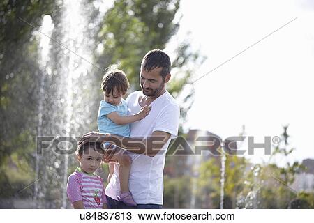 Stock Image - Mid adult man and two daughters standing in water fountains, Madrid, Spain. Fotosearch