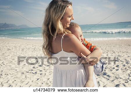 Stock Photo - Mid adult mother carrying young son on beach, Cape Town, Western Cape, South Africa. Fotosearch