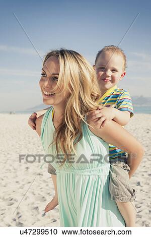 Stock Image - Mid adult mother giving young son piggy back on beach, Cape Town, Western Cape, South Africa. Fotosearch