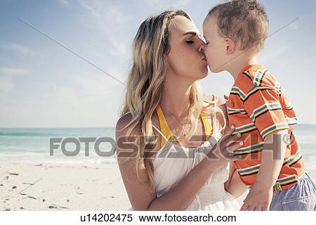 Stock Photography - Mid adult mother kissing young son on beach, Cape Town, Western Cape, South Africa. Fotosearch