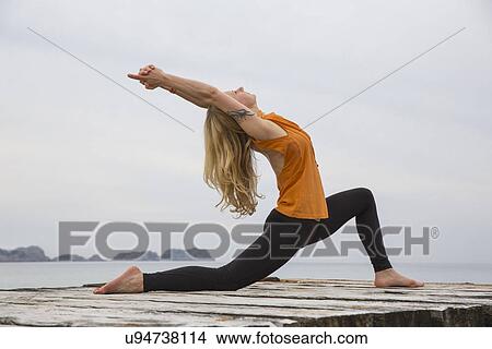 Picture - Mid adult woman bending backwards practicing yoga on wooden sea pier. Fotosearch