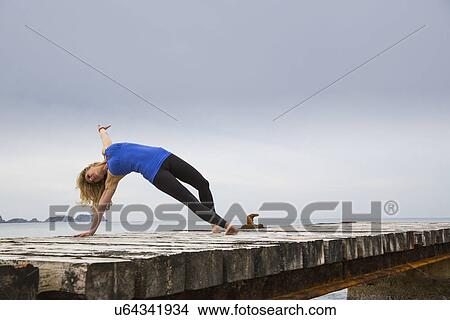 Picture - Mid adult woman practicing yoga move on wooden sea pier. Fotosearch