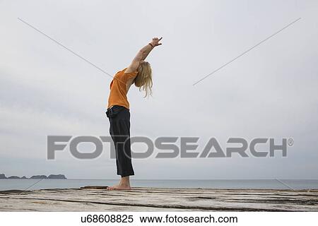 Stock Photography - Mid adult woman stretching back practicing yoga on wooden sea pier. Fotosearch
