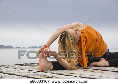 Stock Photo - Mid adult woman touching toes practicing yoga on wooden sea pier. Fotosearch
