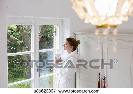 Stock Photo - Mid adult woman wearing bathrobe looking out of window. Fotosearch