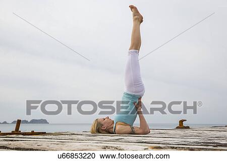Stock Image - Mid adult woman with legs raised practicing yoga on wooden sea pier. Fotosearch