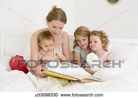 Stock Image - Mother reading book to three daughters in bed. Fotosearch