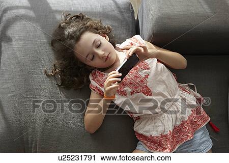 Overhead view of girl lying on living room sofa playing with smartphone View Large Photo Image Stock Image - Overhead view of girl lying on living room sofa playing with smartphone. Fotosearch