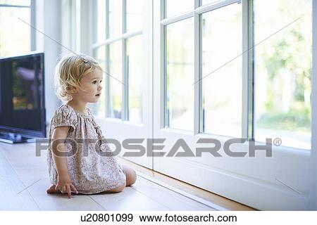 Stock Photo - Portrait female toddler sitting on floor looking out of patio doors. Fotosearch