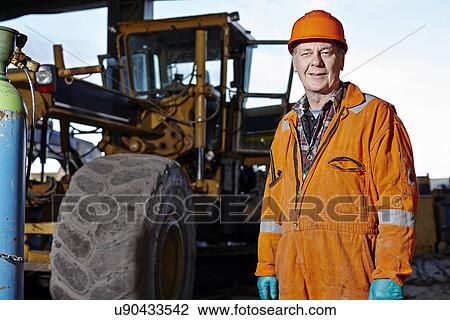 Stock Image - Portrait of quarry worker next to excavator at quarry site. Fotosearch