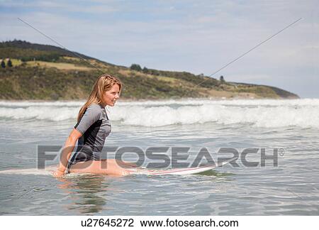 Stock Image - Portrait of young woman sitting on surfboard in sea. Fotosearch