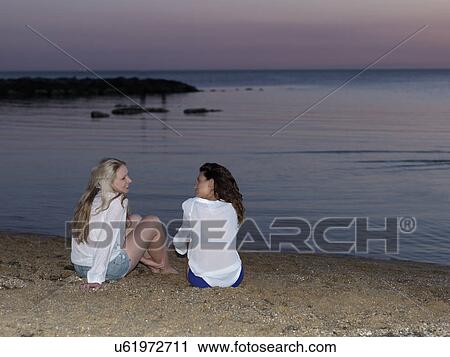 Stock Image - Rear view of two young women friends sitting chatting on beach at sunset, Williamstown, Melbourne, Australia. Fotosearch