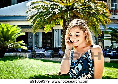Stock Photo - Smiling young woman chatting on smartphone in city park. Fotosearch