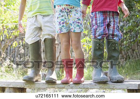 Stock Image - Three children on garden seat in over sized rubber boots. Fotosearch