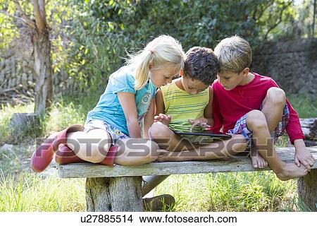 Three children sitting on garden seat looking down at digital tablet View Large Photo Image Picture - Three children sitting on garden seat looking down at digital tablet. Fotosearch