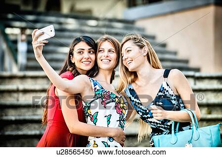 Stock Image - Three fashionable young women taking selfie on stairway, Cagliari, Sardinia, Italy. Fotosearch