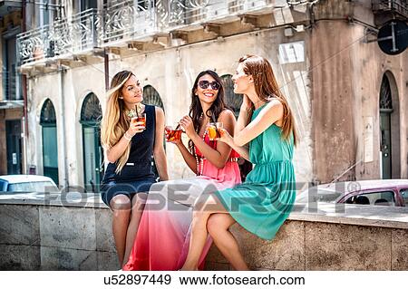 Stock Photo - Three young fashionable female friends having cocktails on sidewalk cafe wall, Cagliari, Sardinia, Italy. Fotosearch
