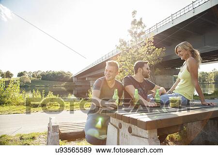 Stock Photo - Three young friends drinking beer on riverside picnic bench. Fotosearch