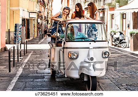Stock Image - Three young women standing up in open back seat of Italian taxi, Cagliari, Sardinia, Italy. Fotosearch