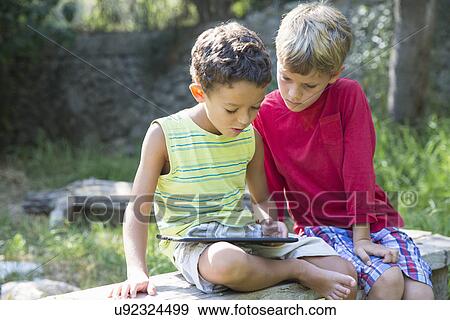 Two boys sitting on garden seat looking down at digital tablet View Large Photo Image Stock Photo - Two boys sitting on garden seat looking down at digital tablet. Fotosearch