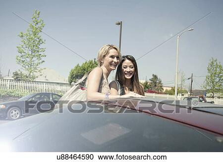 Stock Image - Two women, laughing, standing beside car. Fotosearch