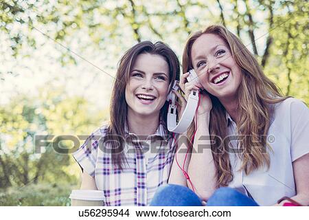 Two young female friends drinking coffee and sharing music on headphones in park View Large Photo Image Picture - Two young female friends drinking coffee and sharing music on headphones in park. Fotosearch