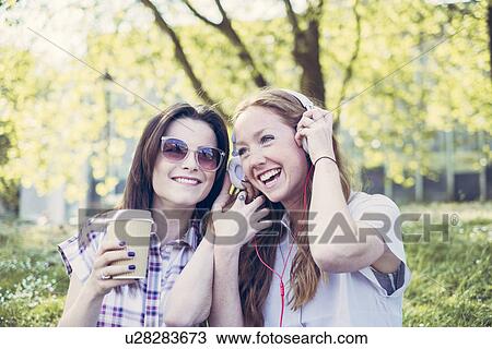 Two young female friends drinking coffee and listening to music on headphones in park View Large Photo Image Stock Image - Two young female friends drinking coffee and listening to music on headphones in park. Fotosearch