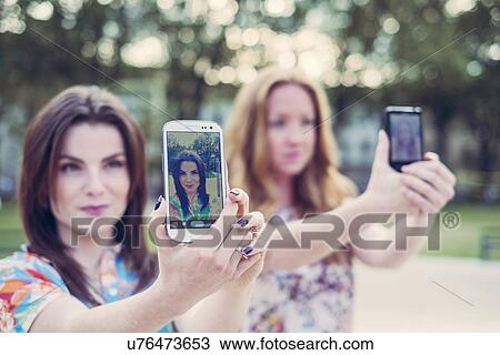 Stock Image - Two young female friends taking selfie on smartphones in unison. Fotosearch