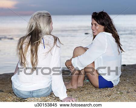 Stock Photography - Two young women friends sitting chatting on beach at sunset, Williamstown, Melbourne, Australia. Fotosearch