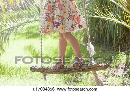 Stock Photograph - Waist down cropped shot of girl standing on tree swing in garden. Fotosearch