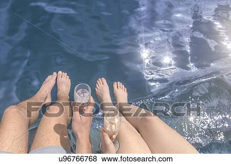 Stock Photo - Young barefoot couple holding champagne glasses over sunlit lake, overhead view. Fotosearch