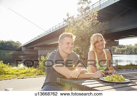 Stock Image - Young couple drinking beer on riverside picnic bench. Fotosearch