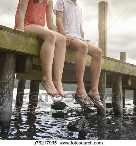 Stock Photography - Young couple holding hands together and sitting on edge of jetty over lake. Fotosearch