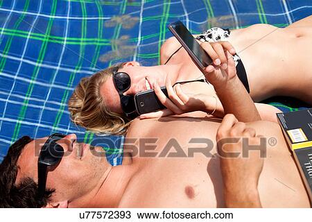 Stock Image - Young couple lying on beach towel using smartphones. Fotosearch