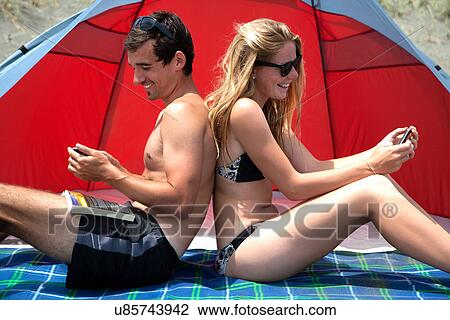 Stock Image - Young couple sitting back to back on beach towel, using smartphones. Fotosearch