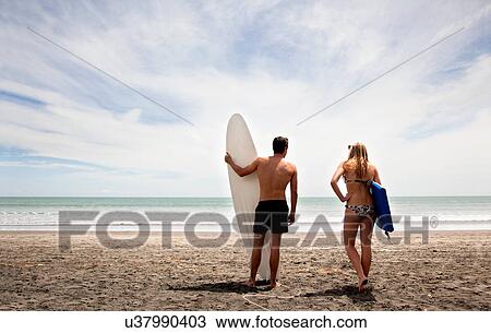 Stock Image - Young couple standing on beach holding surfboard and boogie board. Fotosearch