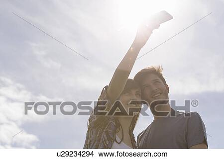 Picture - Young couple taking self portrait on mobile phone under bright sunny sky. Fotosearch