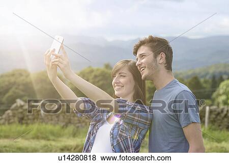 Stock Photo - Young couple taking self portrait on mobile phone in countryside under sunny sky. Fotosearch