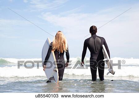 Stock Image - Young couple walking out to sea holding surfboards, rear view. Fotosearch