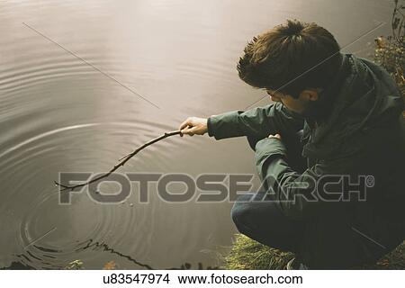 Young man crouching on riverbank making ripples in water with twig View Large Photo Image Picture - Young man crouching on riverbank making ripples in water with twig. Fotosearch