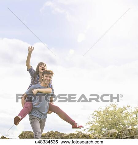 Stock Photography - Young man giving piggyback ride to girlfriend in countryside under bright sunny sky. Fotosearch