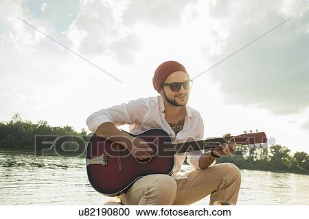 Stock Image - Young man sitting by lake playing guitar. Fotosearch