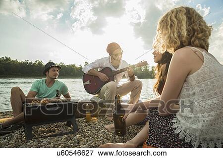 Stock Photo - Young man sitting by lake with friends playing guitar. Fotosearch