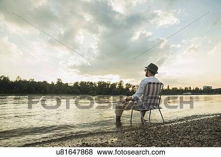 Young man sitting on chair by lakeside View Large Photo Image Stock Photo - Young man sitting on chair by lakeside. Fotosearch
