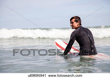 Stock Image - Young man sitting on surfboard in sea. Fotosearch