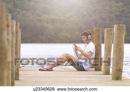 Young man wearing headphones and using mp3 player on jetty over lake View Large Photo Image Stock Photo - Young man wearing headphones and using mp3 player on jetty over lake. Fotosearch
